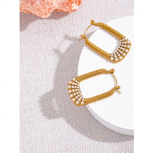 A pair of gold-toned hoop earrings with a textured, ribbed band and a curved row of small clear stones on the front, displayed on a white round pedestal over a white textured surface.